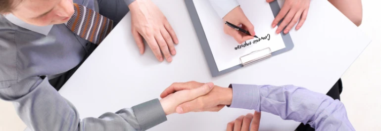  Dos personas se dan la mano sobre una mesa con un documento y una persona escribiendo. 