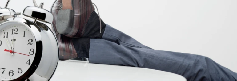  Un reloj despertador grande frente a una persona descansando con zapatos de vestir sobre una superficie. 