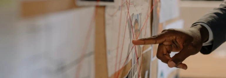  mano señalando con el dedo a un corcho con fotos, apuntes e hilos rojos 