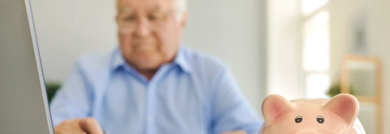 Un hombre mayor trabajando en una computadora con una alcancía de cerdo en primer plano. 