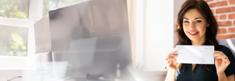 Una mujer sonriente sostiene un cheque en una oficina con luz natural. 