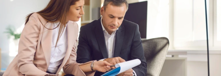  mujer con traje salmón sentada junto a hombre con traje oscuro revisando un documento 