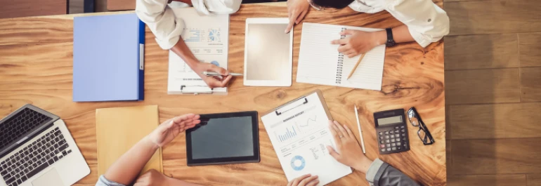  Cuatro personas trabajando juntas en una mesa con laptops, tabletas, calculadoras y documentos. 