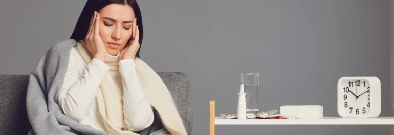  Una mujer enferma se cubre con una manta y se toca la cabeza mientras hay medicinas y un vaso de agua sobre una mesa al lado. 