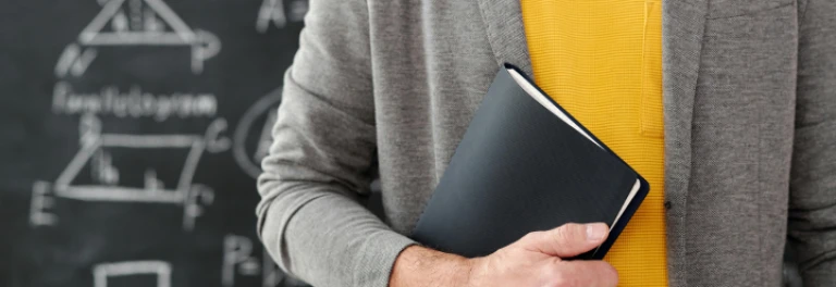  Persona sosteniendo libro negro con jersey gris y camiseta amarilla, frente a pizarra 