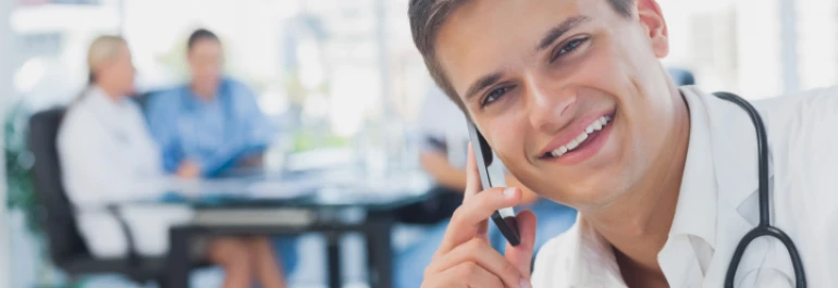  Un médico sonriente habla por teléfono mientras otras personas trabajan en el fondo. 