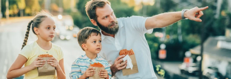  Un hombre y dos niños están comiendo en la calle mientras él señala algo a la distancia. 