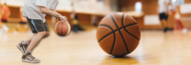  Una pelota de baloncesto en el suelo de una cancha y una persona driblando en el fondo. 