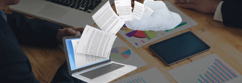  Documentos flotan desde una computadora portátil a una nube sobre una mesa con gráficas y una tablet. 