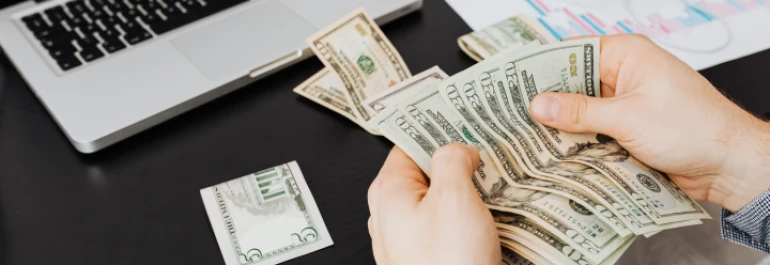  Una persona cuenta billetes de dólares junto a una computadora portátil. 