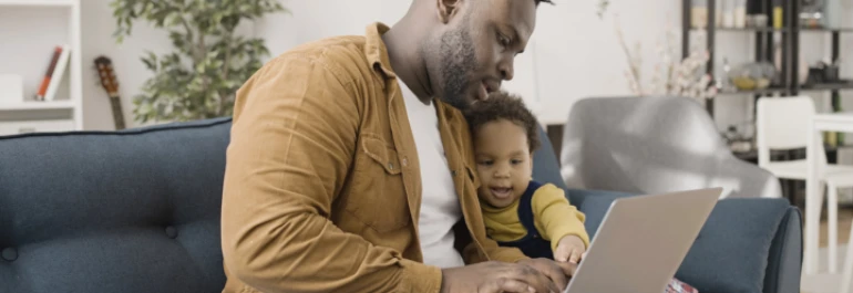  Un hombre sentado en un sofá utiliza una laptop junto a un niño pequeño. 