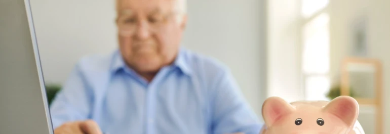  Un hombre mayor está usando una computadora portátil cerca de una alcancía de cerdito. 