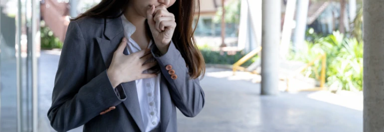  Mujer de traje tosiendo, sujetándose el pecho con una mano, mientras se cubre la boca con la otra 