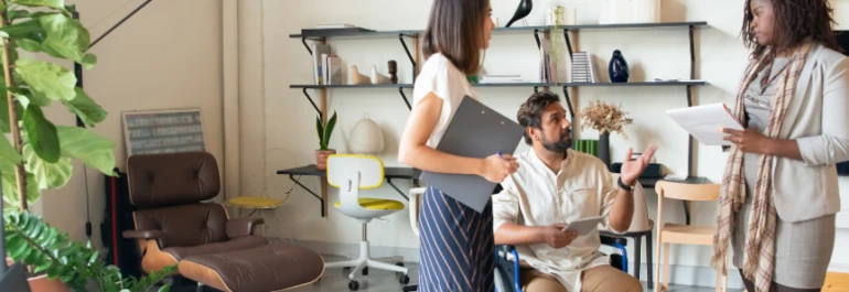  Tres personas dialogan en una oficina moderna con estanterías y una planta. 