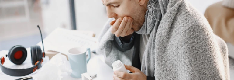  Un hombre cubierto con una manta revisa un frasco de medicamento mientras está sentado junto a una mesa con una taza y audífonos. 