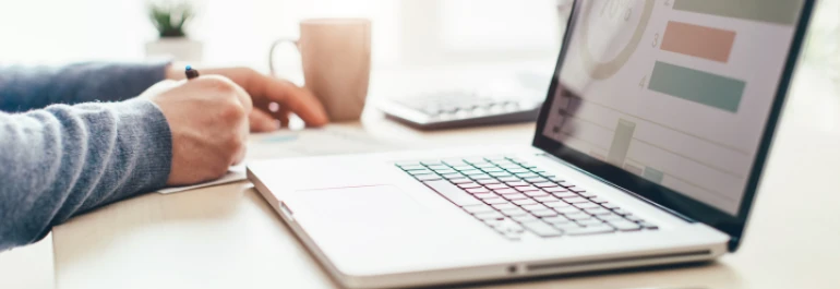  Una persona trabaja en un escritorio con una computadora portátil y toma notas junto a una taza. 