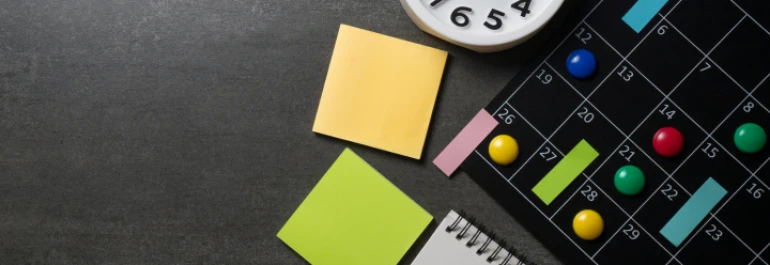  Un calendario con chinchetas de colores, notas adhesivas, un reloj y un bloc de notas sobre una superficie oscura. 