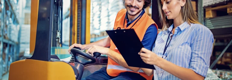  Dos personas revisan un documento en un almacén mientras una de ellas está en un montacargas. 