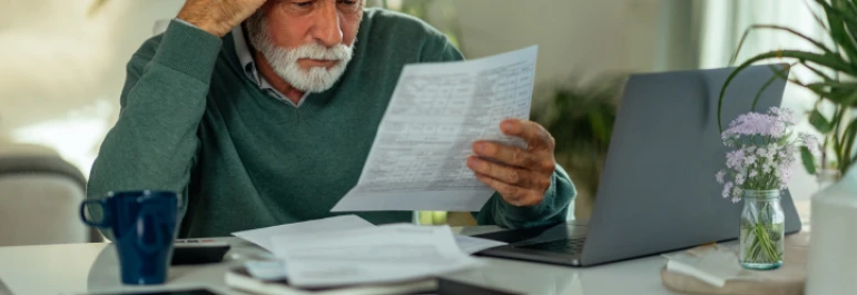  Un hombre mayor revisa preocupado unos documentos frente a una computadora portátil. 