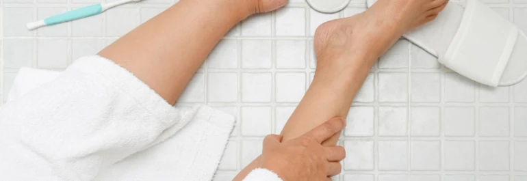  Una persona en bata de baño está sentada en el suelo de cerámica con una pierna desnuda hacia adelante, junto a un cepillo dental y una zapatilla. 