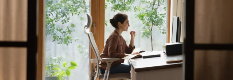  Una persona está trabajando en una computadora en un espacio hogareño luminoso con ventanales y plantas. 