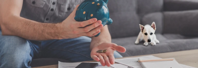  Un hombre está sacando monedas de una alcancía mientras un perro lo observa desde el sofá. 