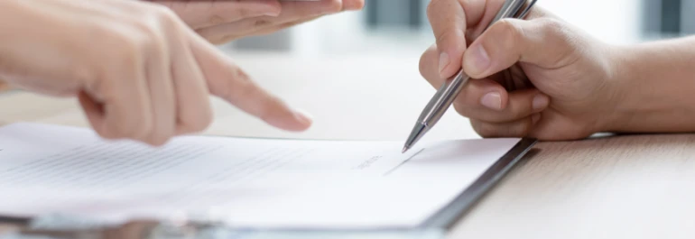  Una mano sostiene un bolígrafo mientras otra señala un documento sobre una mesa. 