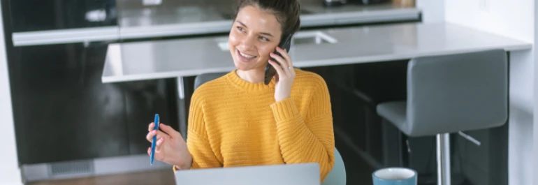  Una persona sonriente con suéter amarillo habla por teléfono mientras usa una computadora portátil en la cocina. 
