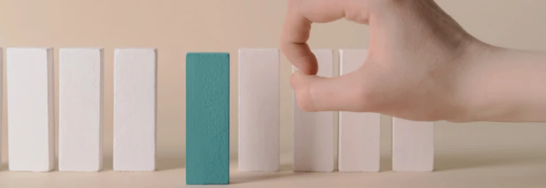  Una mano detiene el derribo de fichas de dominó, destacando una diferente de color azul entre las blancas. 