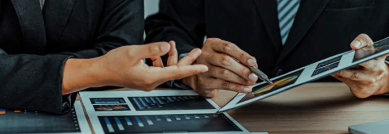  Dos personas revisan gráficos en documentos y una tablet sobre una mesa. 