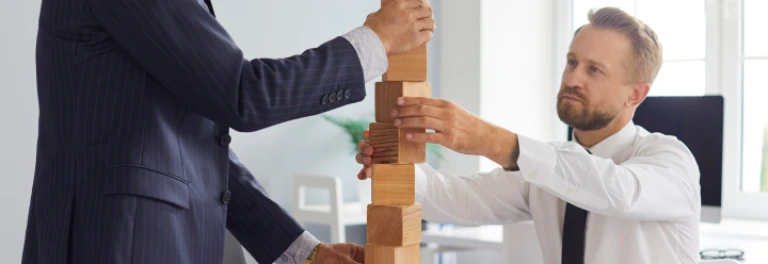  Dos hombres de traje y corbata colocando una inestable torre de bloques de madera en una oficina 