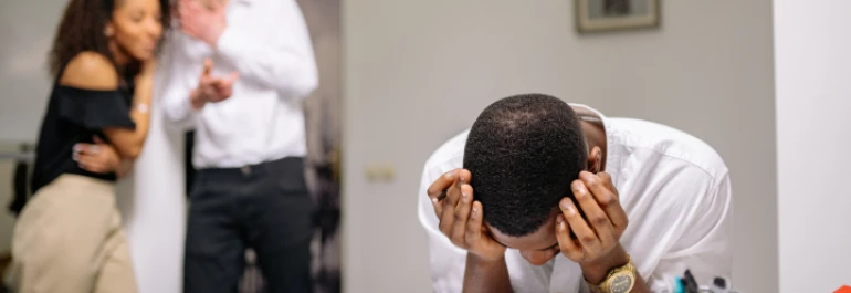  Un hombre se cubre la cara con las manos mientras dos personas hablan en el fondo. 