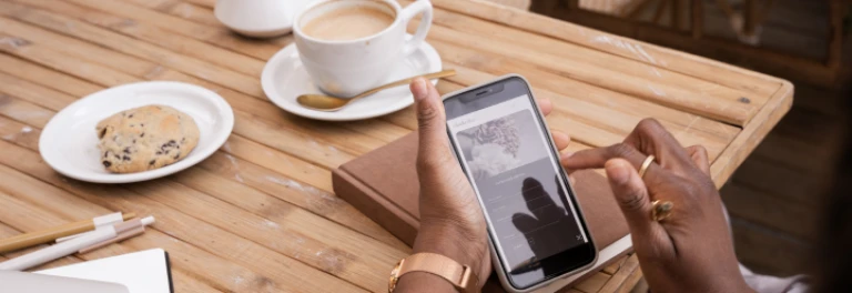  Manos sosteniendo el móvil en la mesa con una galleta, un café y un cuaderno 