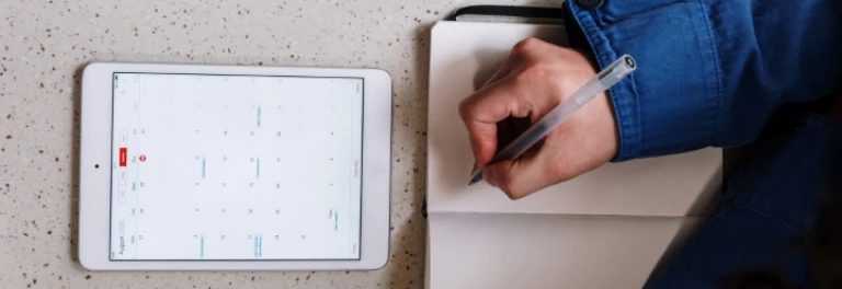  iPad sobre escritorio con el calendario abierto junto a una mano escribiendo sobre una libreta 