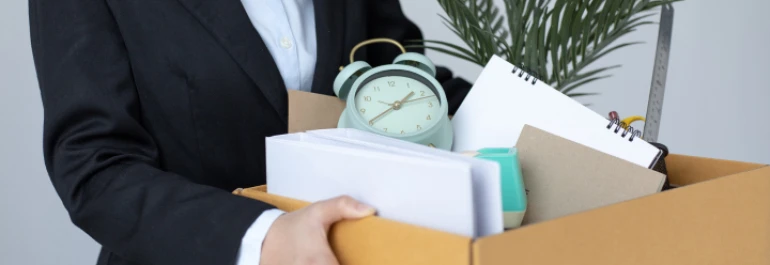  Una persona lleva una caja con objetos de oficina, incluyendo un reloj, cuadernos y una planta. 