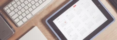  iPad con calendario abierto frente a teclado, junto a cuaderno y iPhone sobre escritorio de madera 