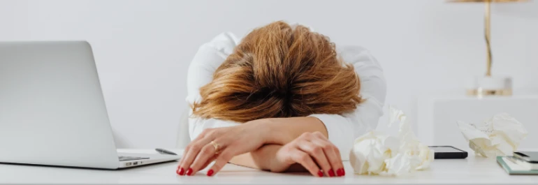  Mujer apoyada sobre sus brazos en un escritorio blanco junto a su laptop y papeles arrugados 