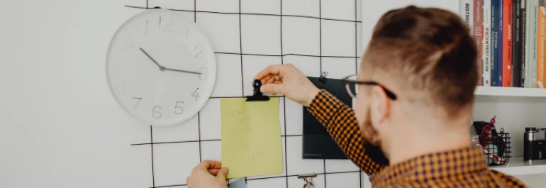  Un hombre está sujetando una nota amarilla con un clip frente a un reloj de pared. 